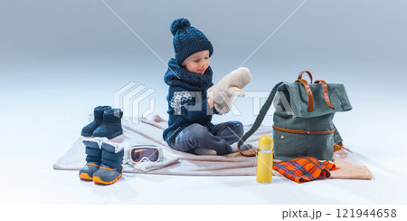 Junior explorer, little boy dressed warm clothes, sitting on blanket and wearing gloves against grey background, ready to adventures. 121944658