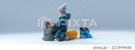 Banner. Curious little boy, dressed warm clothes, sitting on luggage ready to rest against grey studio background with negative space to insert text. 121944680
