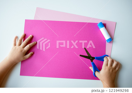 Childs hands crafting with pink paper, blue scissors, and glue on light background Childs hands crafting with pink paper, blue scissors, and glue on light background 121949298