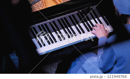 Close-up of piano keys with a person's hand in a blue sleeve touching the keyboard, showing part of the black grand piano body and tuning pins in dramatic lighting. Top view 121949768