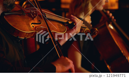 A close-up of musicians playing string instruments in an orchestra, focusing on a violinist and a cellist. The warm lighting highlights the wood of the instruments 121949771