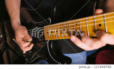 A close-up shot of a person playing a black electric guitar. The focus is on the hands, one holding a green pick and the other pressing the strings on the fretboard A close-up shot of a person playing a black electric guitar. The focus is on the hands, one holding a green pick and the other pressing the strings on the fretboard 121949789