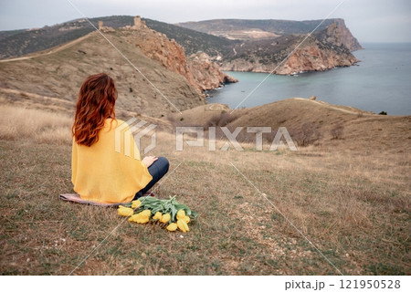 A woman sits on the grass with a bouquet of yellow flowers in her lap. She is looking out at the ocean, taking in the view. Concept of peace and tranquility. 121950528
