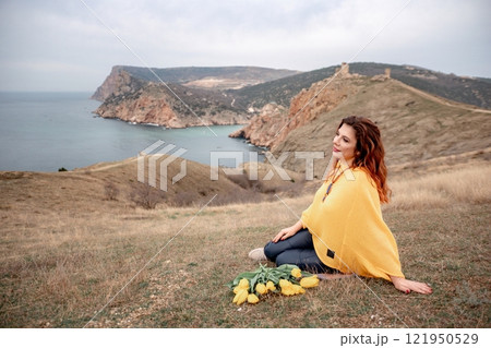 A woman sits on the grass with a bouquet of yellow flowers in her lap. She is looking out at the ocean, taking in the view. Concept of peace and tranquility. 121950529