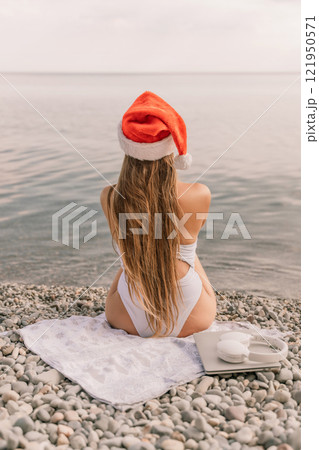A woman wearing a red Santa hat sits on a beach with her legs crossed. She is wearing a white bikini and has a laptop on her lap. The beach is rocky and the water is calm. 121950571