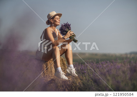 woman sitting in a field of lavender and wearing a straw hat. She is smiling and holding a bouquet of flowers. Scene is peaceful and serene, as the woman is surrounded by the beauty of nature 121950584