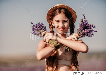 girl is holding a bunch of lavender purple flowers in her hands and wearing a straw hat. She is smiling and she is enjoying herself. The scene is set in a field of lavender, which adds to the peaceful 121950585