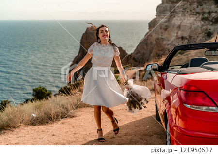 A woman in a white dress is standing next to a red convertible car. She is holding a bouquet of flowers and she is in a happy mood. The scene suggests a romantic or special occasion. 121950617