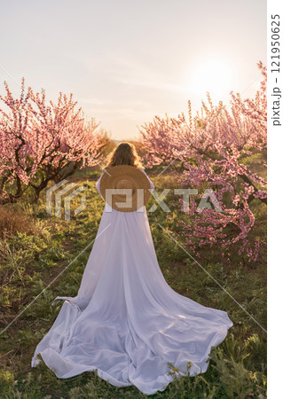Woman blooming peach orchard. Against the backdrop of a picturesque peach orchard, a woman in a long white dress and hat enjoys a peaceful walk in the park, surrounded by the beauty of nature. Woman blooming peach orchard. Against the backdrop of a picturesque peach orchard, a woman in a long white dress and hat enjoys a peaceful walk in the park, surrounded by the beauty of nature. 121950625