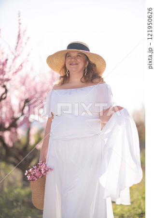 Woman blooming peach orchard. Against the backdrop of a picturesque peach orchard, a woman in a long white dress and hat enjoys a peaceful walk in the park, surrounded by the beauty of nature. Woman blooming peach orchard. Against the backdrop of a picturesque peach orchard, a woman in a long white dress and hat enjoys a peaceful walk in the park, surrounded by the beauty of nature. 121950626
