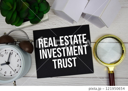 Real Estate Investment Trust text message on sticky note with alarm clock, magnifying glass and house model on wooden backgroun Real Estate Investment Trust text message on sticky note with alarm clock, magnifying glass and house model on wooden backgroun 121950654