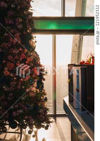 A Christmas tree with pink and gold ornaments stands by a large window, overlooking the London cityscape. The setting sun casts a warm glow inside. A Christmas tree with pink and gold ornaments stands by a large window, overlooking the London cityscape. The setting sun casts a warm glow inside. 121952312