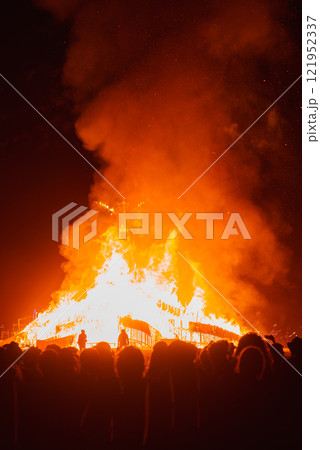 A towering bonfire illuminates the night sky, surrounded by silhouetted spectators. Sparks fly as the flames cast an orange glow in the Black Rock Desert. 121952337