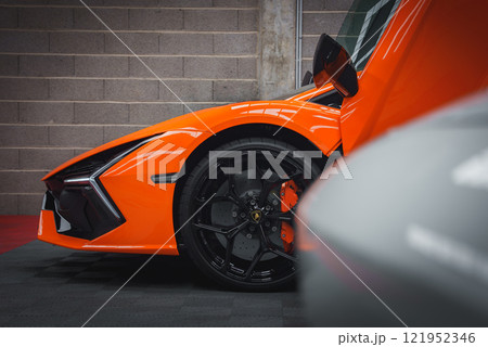 The image captures a close up of an orange Lamborghini Revuelto, focusing on the front wheel, black alloy wheels, and orange brake calipers against a concrete wall. 121952346