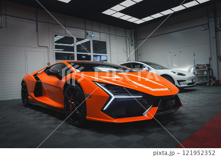 An orange Lamborghini Revuelto is parked indoors, showcasing its sleek design and LED headlights. A checkered floor and grid ceiling enhance the setting. 121952352