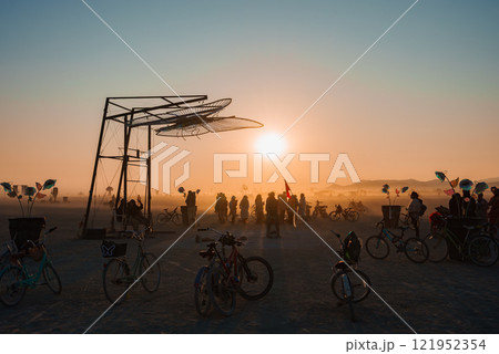 Participants gather around a large metal sculpture at a desert festival, with bicycles and flags. The setting sun casts a warm glow over the dusty landscape. 121952354