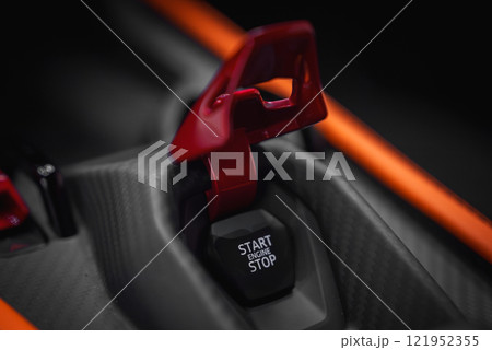Detailed view of the Lamborghini Revuelto's start or stop engine button with a red safety cover, set in a carbon fiber panel with orange accents. 121952355