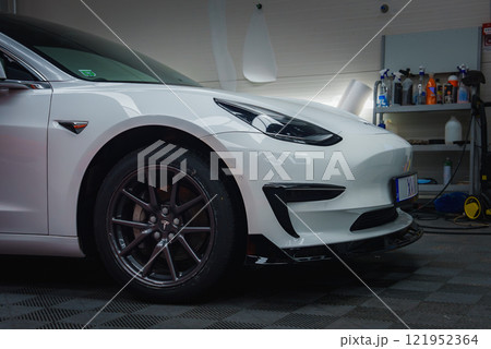 A white Tesla vehicle is shown indoors, focusing on the front left side with wheel and headlight. Tiled floor and shelves with tools are visible in the background. A white Tesla vehicle is shown indoors, focusing on the front left side with wheel and headlight. Tiled floor and shelves with tools are visible in the background. 121952364