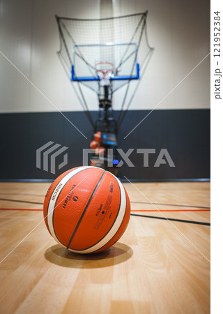 A basketball rests on a polished wooden court with a hoop and net in the background, featuring a ball return system in a modern indoor facility. 121952384