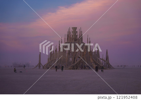 A large wooden dome with spires and latticework stands in the desert. The sky is pink and purple, and participants walk around the installation. 121952408