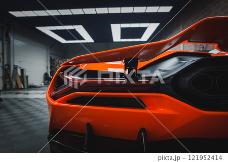 The image captures the rear section of a Lamborghini Revuelto, focusing on its taillights and rear diffuser. The vibrant orange car is in a modern showroom. 121952414