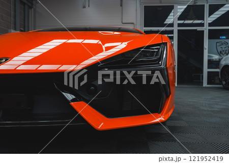 The image shows a close up of a Lamborghini Revuelto's front end, highlighting its angular design, vibrant orange color, and unique headlights indoors. 121952419