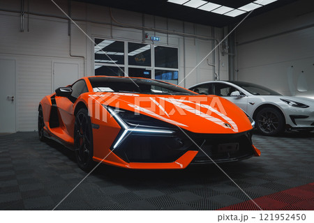 An orange Lamborghini Revuelto is parked indoors on a checkered floor, showcasing its angular headlights and aerodynamic body. A white car is beside it. 121952450