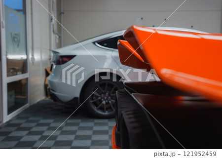 The image shows a close up of a Lamborghini Revuelto's vibrant orange rear wing in an indoor showroom, with tiled flooring and a white car nearby. 121952459