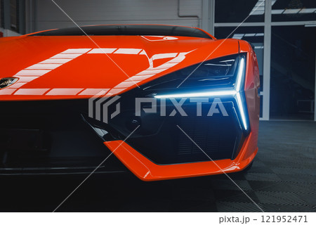 The image captures the front end of a Lamborghini Revuelto indoors, focusing on its angular design, vibrant orange color, and distinctive LED headlight. The image captures the front end of a Lamborghini Revuelto indoors, focusing on its angular design, vibrant orange color, and distinctive LED headlight. 121952471