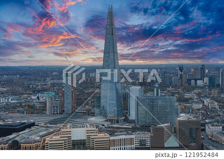 The Shard skyscraper stands against a vibrant sunset sky in London. Modern buildings and railway tracks of London Bridge station are visible below. 121952484
