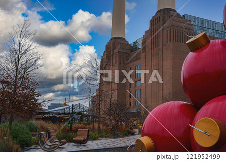 Battersea Power Station's chimneys rise against a partly cloudy sky, with oversized red Christmas ornaments in the foreground, amid winter landscaping. 121952499