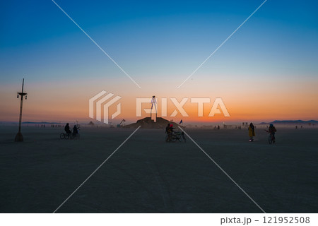 A large wooden art installation stands in a desert at sunset, surrounded by bicyclists. The sky transitions from orange to blue, with mountains in the distance. 121952508