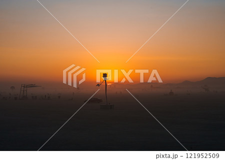 A tall, abstract art installation resembling a human figure is silhouetted against a vibrant sunset in Nevada's Black Rock Desert, with distant mountains visible. 121952509