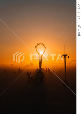 A large human figure sculpture with raised arms is silhouetted against a sunset at a desert festival, surrounded by participants and bicycles. 121952582