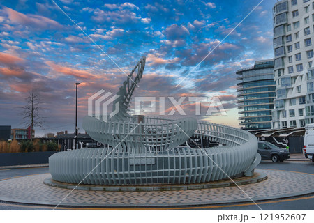 A modern spiral sculpture of white metal slats stands against contemporary buildings in London, with a vibrant sunset sky enhancing the urban scene. A modern spiral sculpture of white metal slats stands against contemporary buildings in London, with a vibrant sunset sky enhancing the urban scene. 121952607