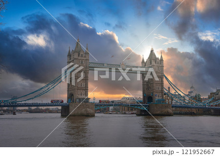 Tower Bridge in London is silhouetted against a vibrant sunset sky. Gothic style towers and suspension elements are visible, with buses crossing above. 121952667