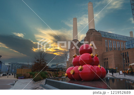 Battersea Power Station's four chimneys stand tall as oversized red Christmas ornaments adorn the foreground, set against a dramatic sunset sky. 121952689