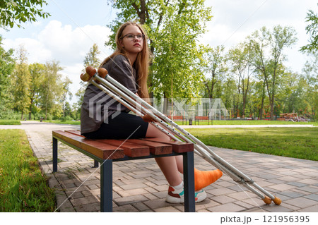 Adolescent girl, perches sullenly on bench, leg adorned in bright orange cast, crutches nearby - her gloomy demeanor contrasts the tranquil summer setting 121956395