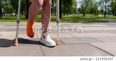 leg encased in an orange cast, with crutches visible in the lush, green background of a summer park setting. Concept of injury recovery leg encased in an orange cast, with crutches visible in the lush, green background of a summer park setting. Concept of injury recovery 121956397