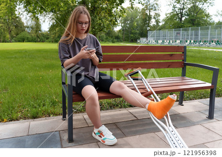 Young female with a casted leg relaxing on a bench in a lush summer park, reading content on her mobile device during her rehabilitation Young female with a casted leg relaxing on a bench in a lush summer park, reading content on her mobile device during her rehabilitation 121956398