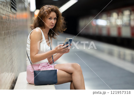Woman looking for subway train schedule on smartphone while sitting on bench at underground metro station 121957318
