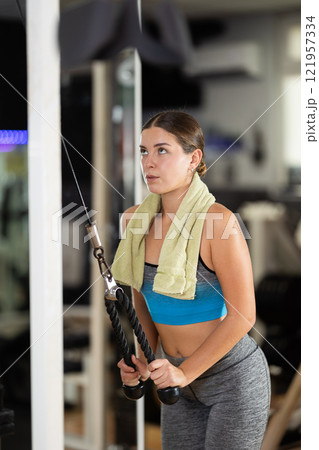 Girl in gym performs exercises for arm muscles using cable crossover machine and pull weighted rope Girl in gym performs exercises for arm muscles using cable crossover machine and pull weighted rope 121957334