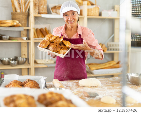 Portrait of old female baker working in bakehouse, holding tray with fresh bakery goods on kitchen Portrait of old female baker working in bakehouse, holding tray with fresh bakery goods on kitchen 121957426