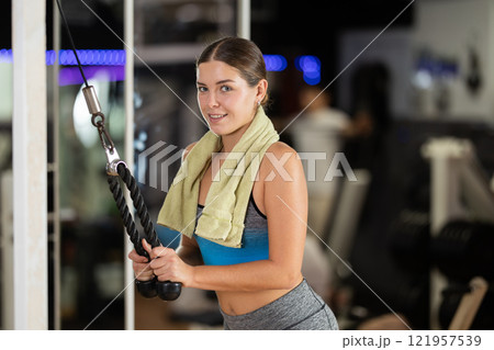 Girl in gym performs exercises for arm muscles using cable crossover machine and pull weighted rope Girl in gym performs exercises for arm muscles using cable crossover machine and pull weighted rope 121957539