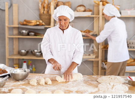 Confidence elderly female baker in white uniform standing at work table and cutting dough into pieces in kitchen of bakery 121957540