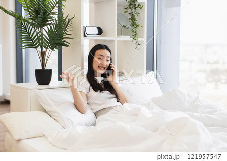 Asian woman in casual white shirt having phone call while relaxing on comfortable bed in bright room. 121957547