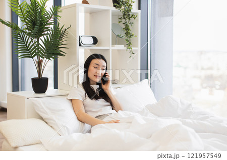 Asian woman in white room sitting on bed talking on phone, creating a calm and relaxed atmosphere. Asian woman in white room sitting on bed talking on phone, creating a calm and relaxed atmosphere. 121957549