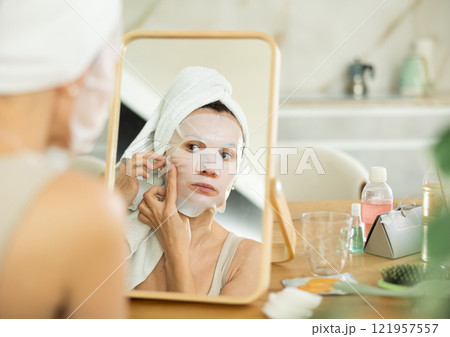Woman applying moisturizing sheet face mask in front of mirror Woman applying moisturizing sheet face mask in front of mirror 121957557