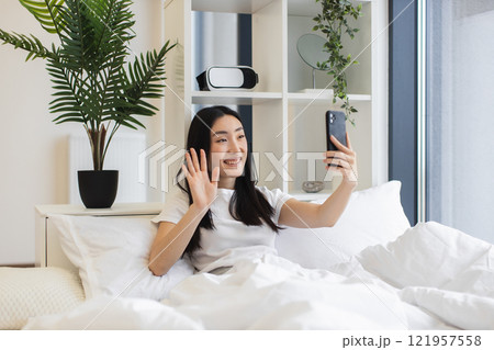 Young Asian woman having online video chat in cozy bedroom setting. She is smiling, holding smartphone, engaging in virtual communication, wearing casual attire. Bright, relaxing indoor environment. 121957558