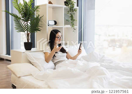 Asian woman in her 20s using smartphone for online video call while sitting on bed. Relaxed atmosphere with indoor plants and modern decor. 121957561